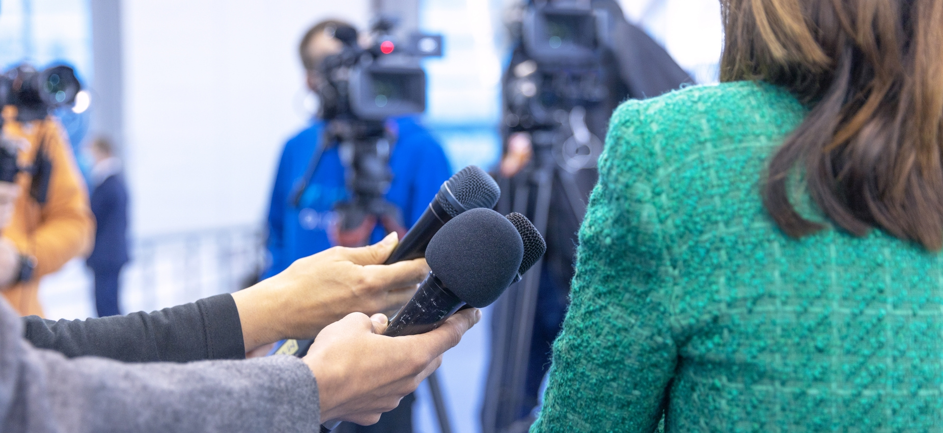 A woman, shown from the back is answering questions to the media. A number of cameras and microphones point in her direction.